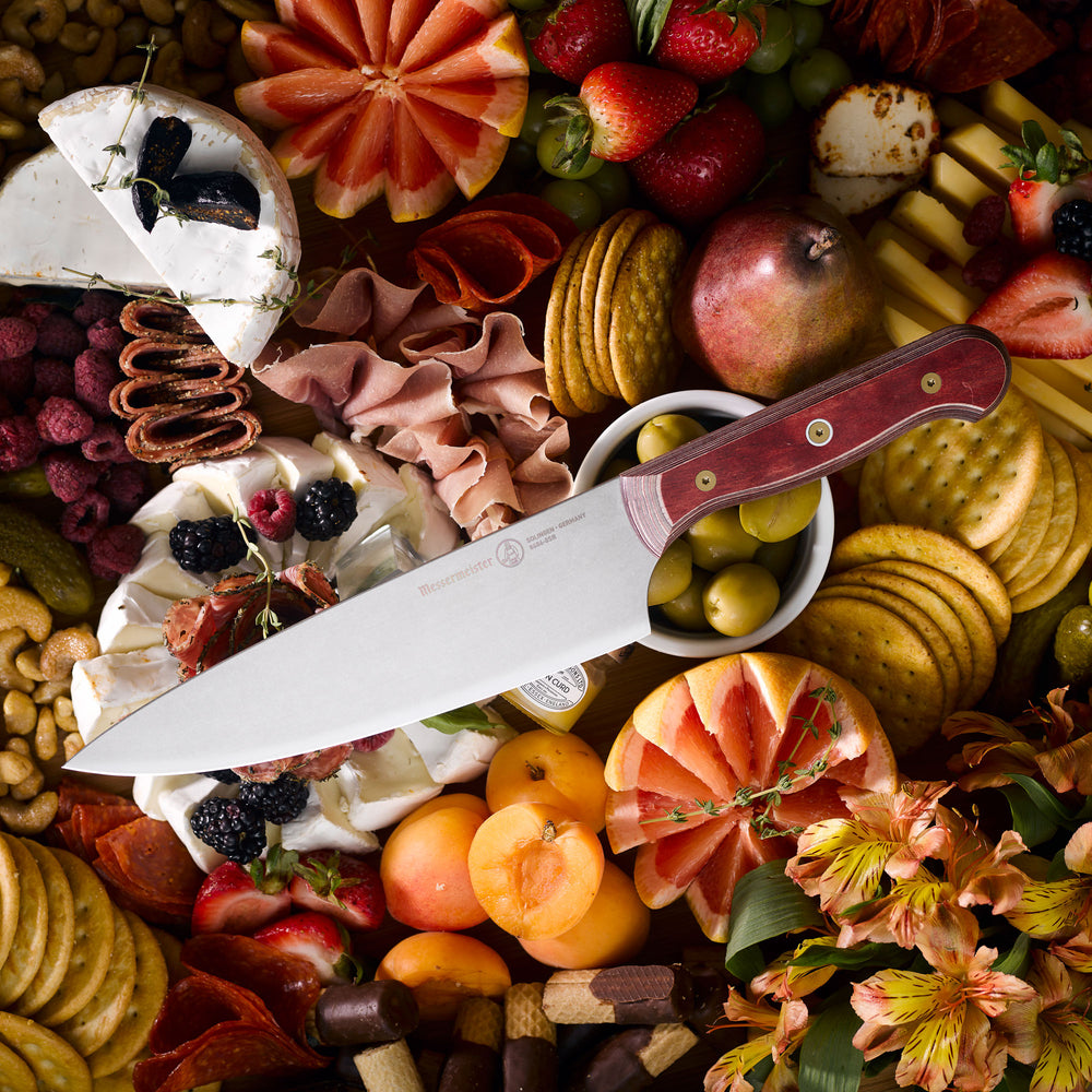 Messermeister Custom Red 8-inch chef’s knife resting on a rustic wooden charcuterie board surrounded by sliced cured meats, artisan cheeses, fresh herbs, and seasonal fruits, highlighting the knife’s satin-finish blade and bold red ergonomic handle in a warm gourmet kitchen setting
