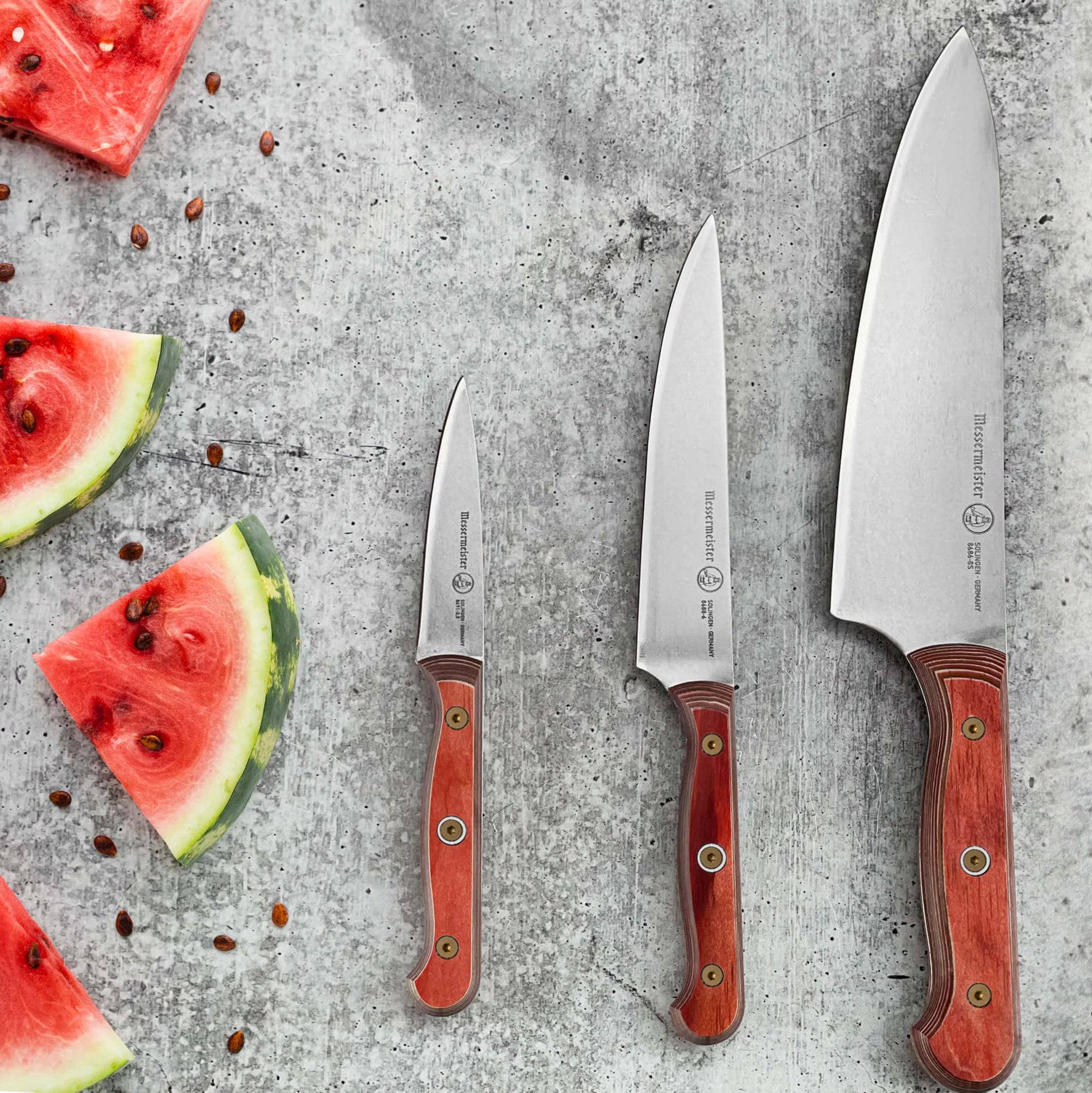 Messermeister Custom 8-inch red chef’s knife alongside a matching paring knife and utility knife, placed near neatly cut watermelon slices that add a fresh pop of color, showcasing the satin stainless steel blades, ergonomic handles, and cohesive modern design