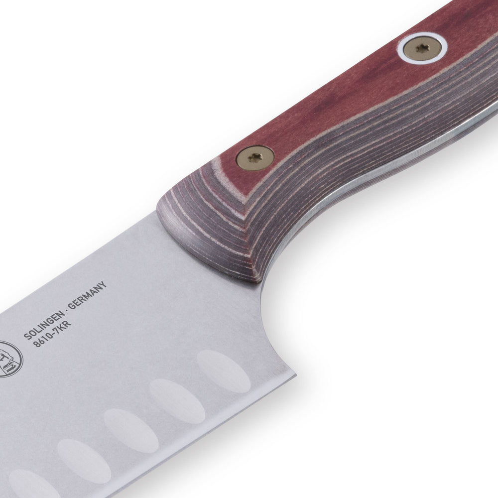 Close-up of the heel of a 7-inch Messermeister Custom Red Kullens Santoku knife, showing the base of the granton-edged stainless steel blade with scalloped hollows meeting the red ergonomic handle.