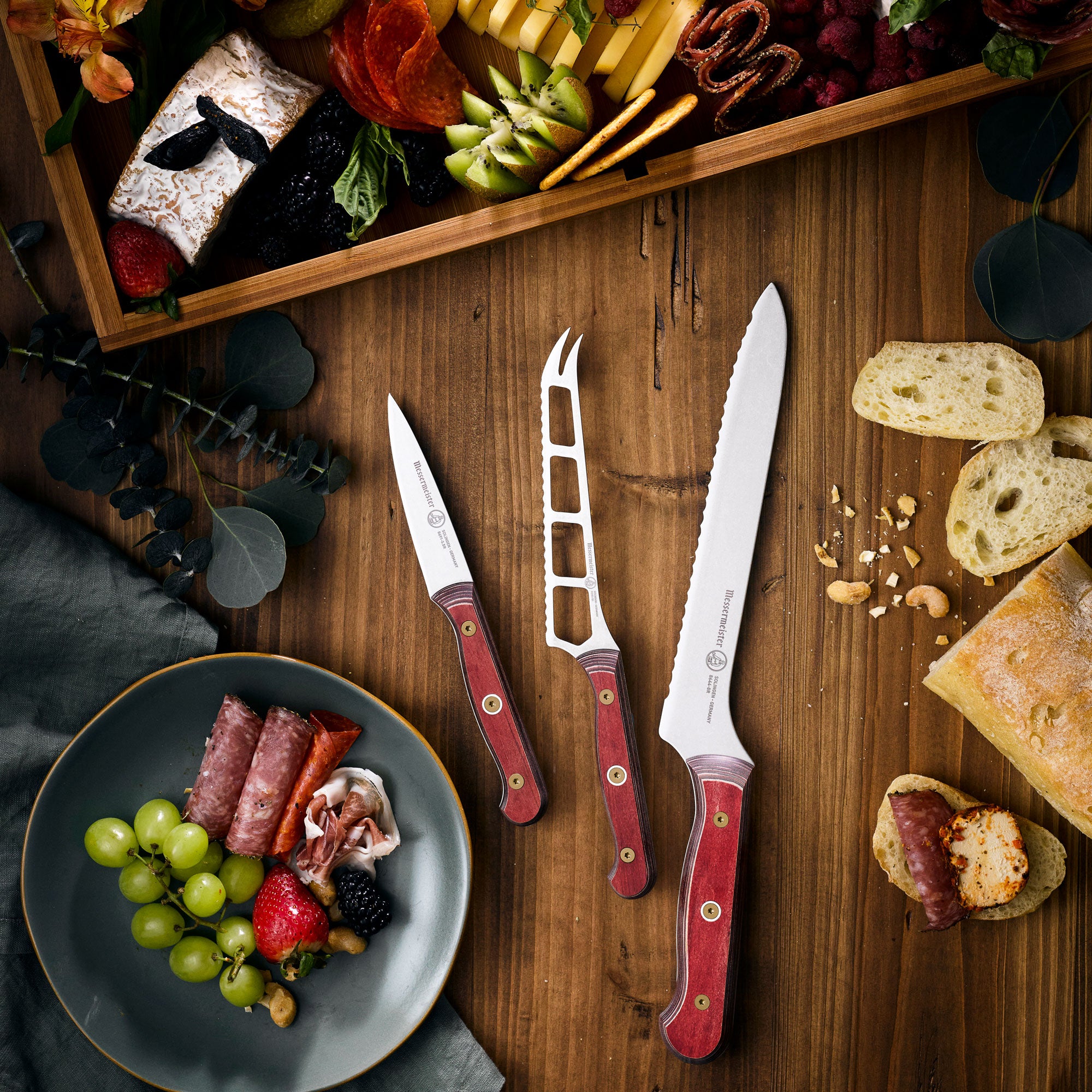  8-inch Messermeister Custom Red scalloped offset knife rests on a wooden cutting board alongside a paring knife, cheese and tomato knife, with a charcuterie spread including sliced meats, cheeses, and fresh tomatoes arranged nearby