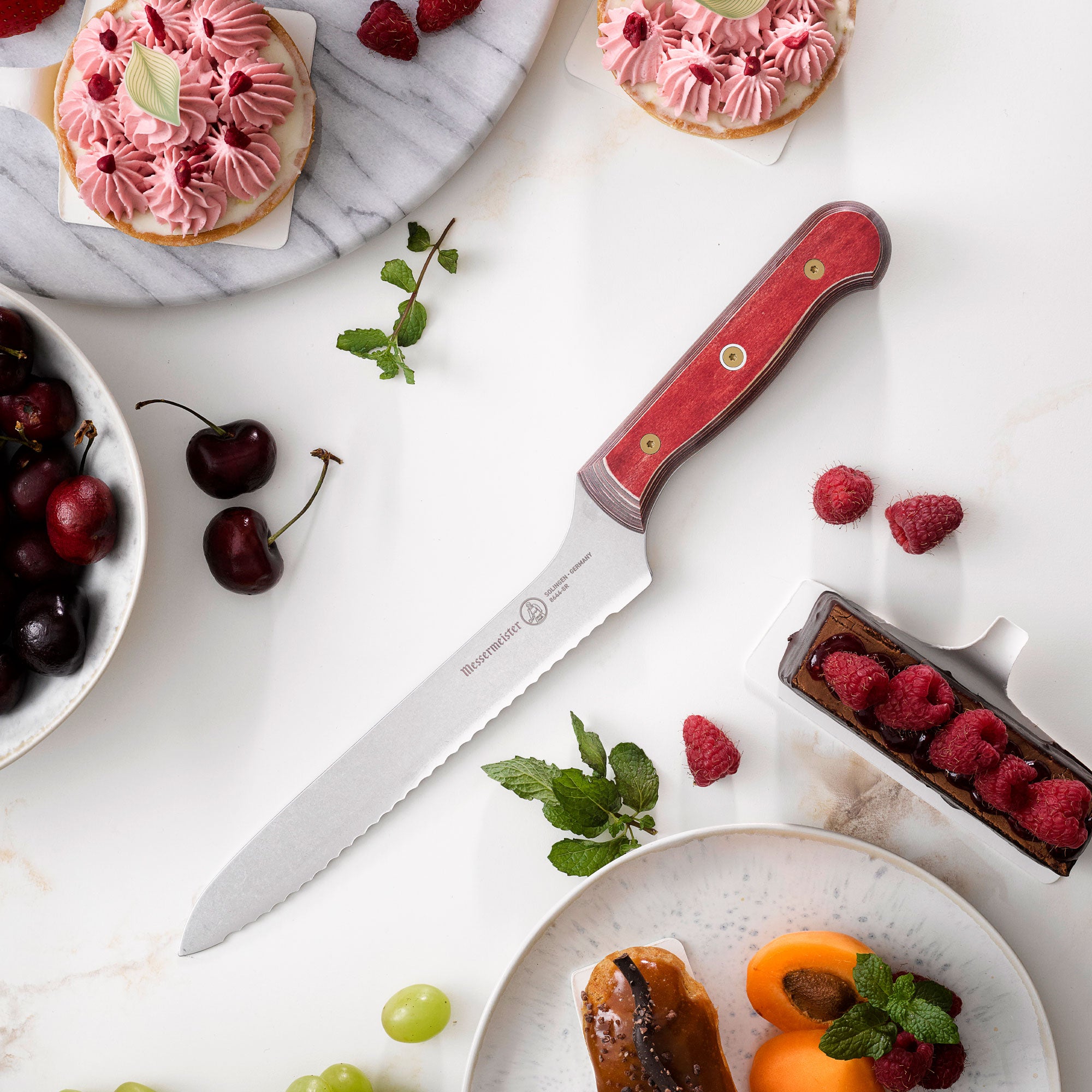 8-inch Messermeister Custom Red scalloped offset knife on white background beside pastries and cherries, showing serrated stainless blade and red ergonomic handle