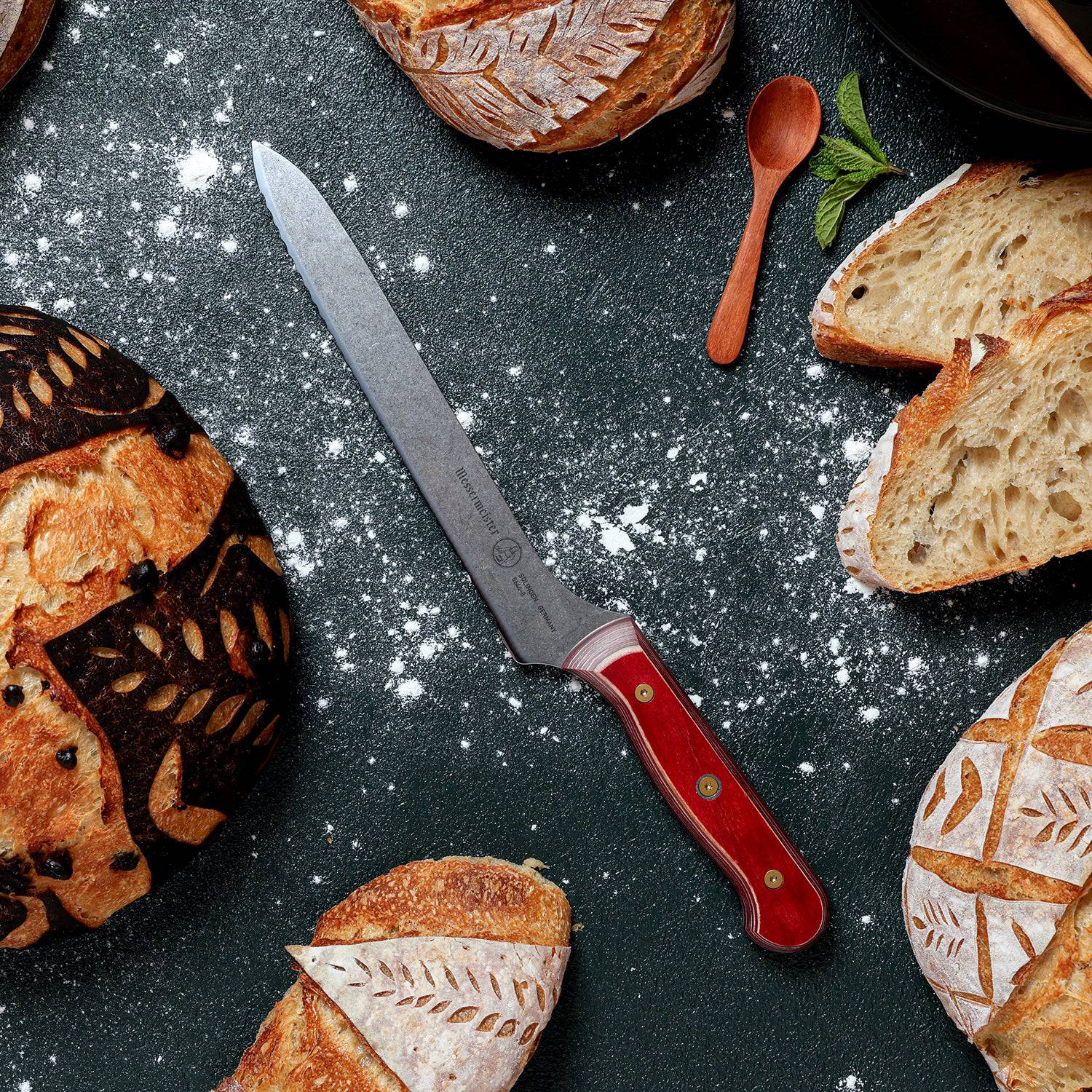 8-inch Messermeister Custom Red scalloped offset knife surrounded by sourdough loaves and sliced bread, highlighting the serrated stainless steel blade and red ergonomic handle