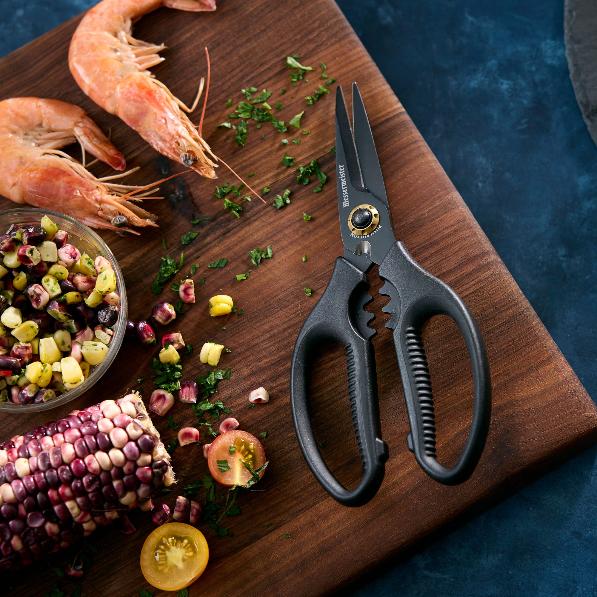 Messermeister Esther Choi Push Button Precision Shears that are all black on a cutting board with prawns, corn and herbs that is laying on a dark blue surface