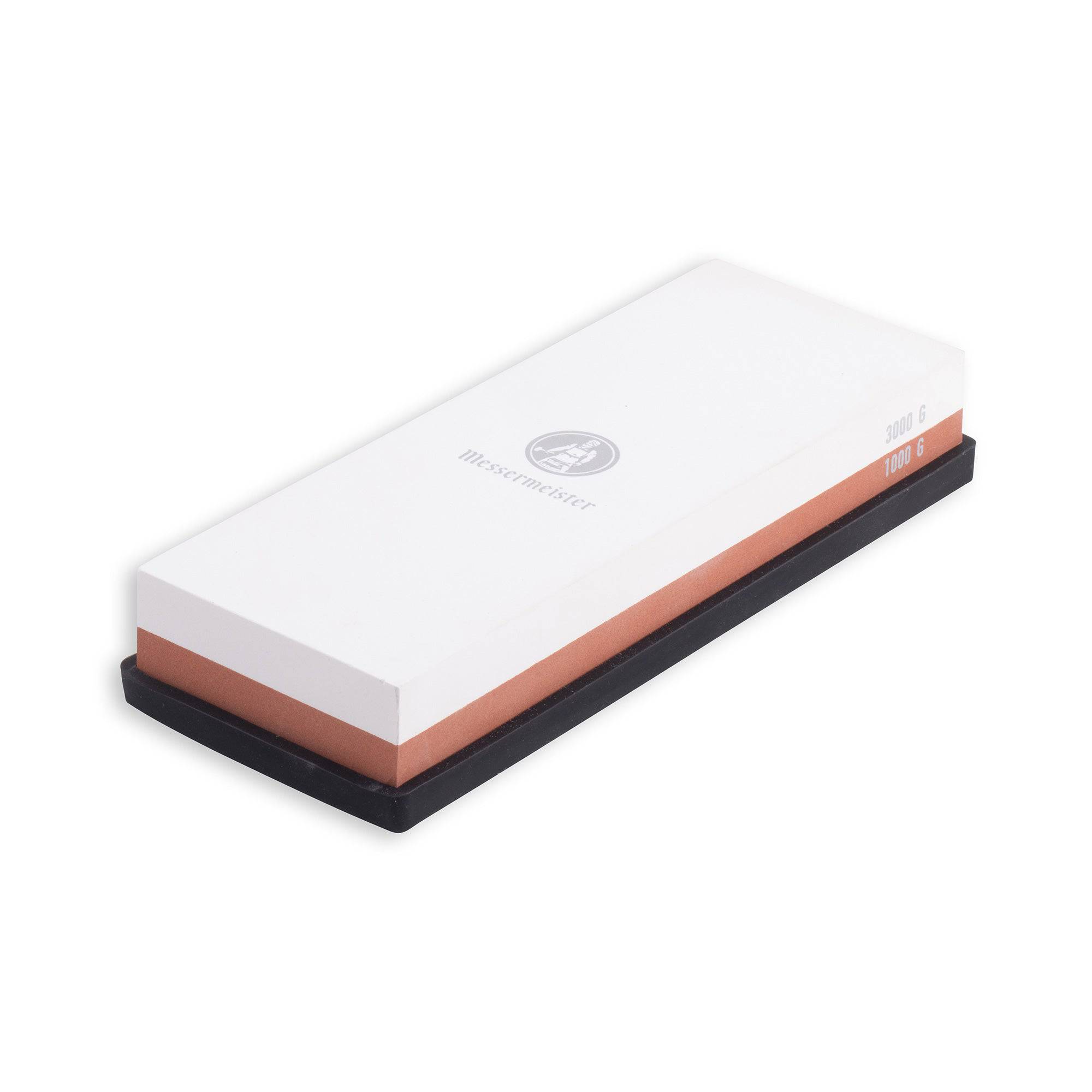 Messermeister  1000 & 3000 Grit Maintenance Stone is a dual sided rectangle 8 inch x 3 inch x 1 inch. The top layer is white with silver "Messermeister" logo.  Bottom layer is reddish brown. The item sits in a rectangular black silicone base. Image tilted slightly up and to the right on a white background.
