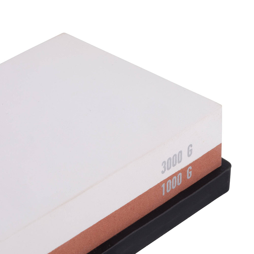 Messermeister  1000 & 3000 Grit Maintenance Stone. shows a close up of the right corner/edge. The top layer is white with silver "3000 G" printed on the edge. Bottom layer is reddish brown with silver "1000 G" printed on edge. The item sits in a black silicone base.