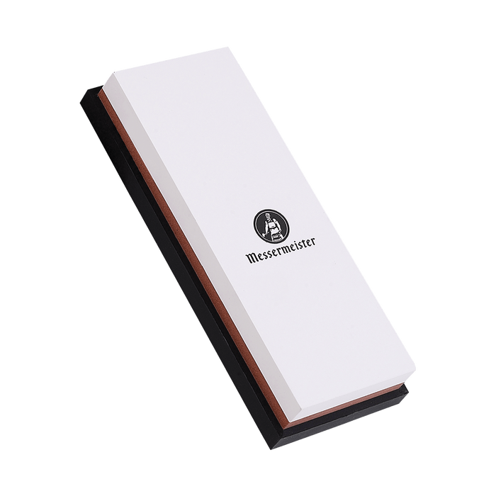 Messermeister 1000 & 3000 Grit Maintenance Stone is a dual sided rectangle 8 inch x 3inch x 1inch. The top layer is white with black "Messermeister" logo.  Bottom layer is reddish. The item sits in a rectangular black silicone base. Image tilted up and to the left.