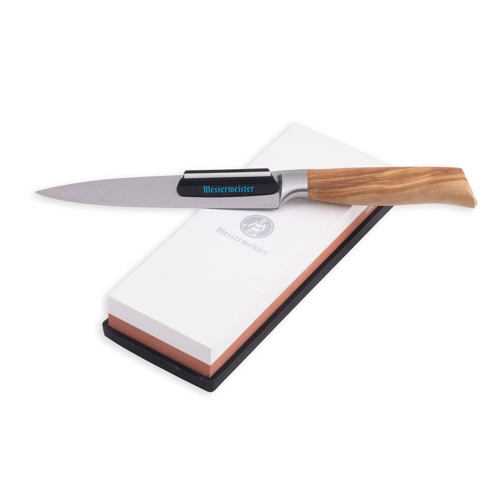 Messermeister 1000 & 3000 grit maintenance Stone  is a dual sided rectangle. The top layer is white with silver "Messermeister" logo.  Bottom layer is reddish brown. The item sits in a rectangular black silicone base. A wooden knife with angle guide is resting along the top of the stone.