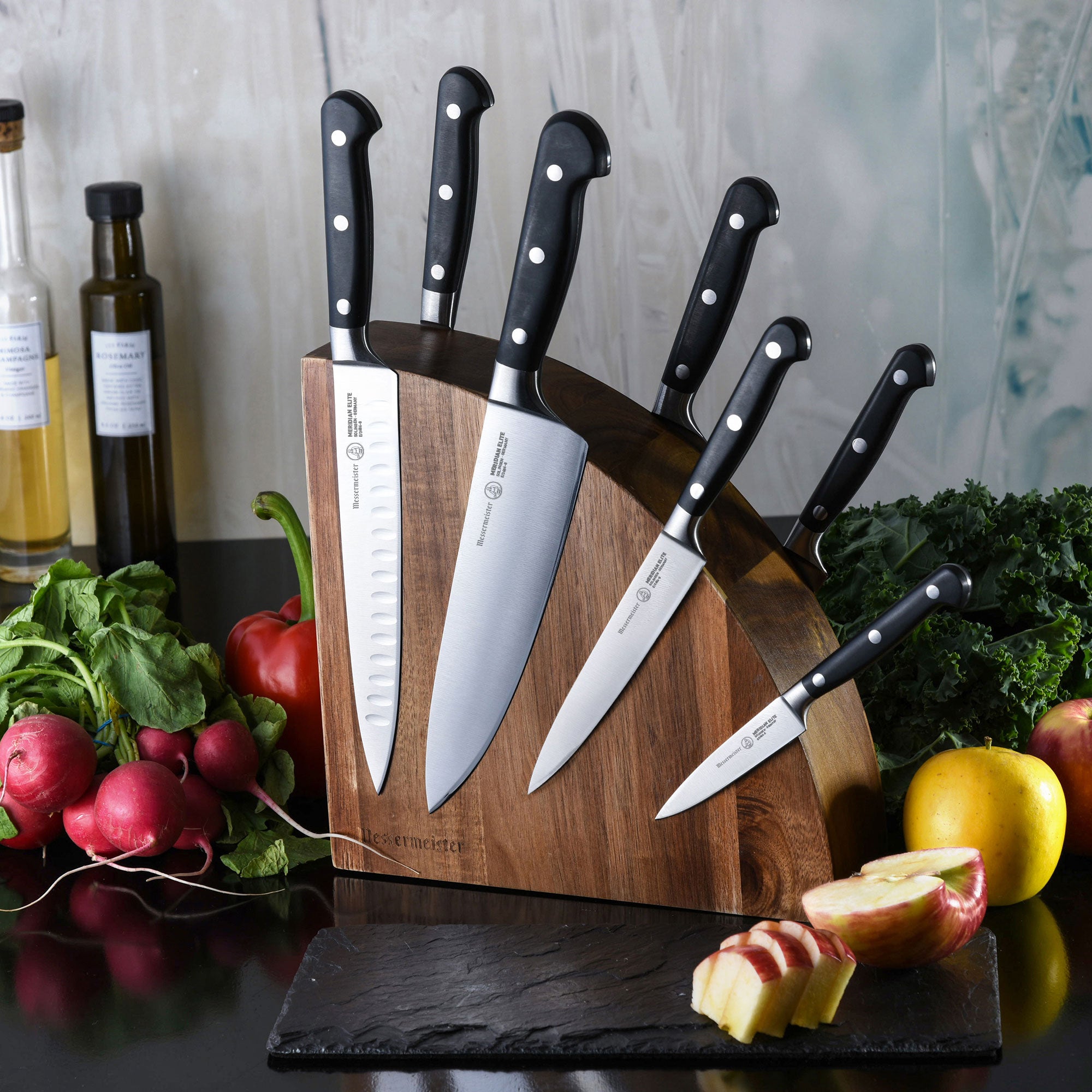 Messermeister Meridian Elite 8 piece Block Set Quarter moon magnetic wooden block with 7 knives attached all with triple rivet black handles. Blades are exposed on the block surface in an arc. Fruits and vegetables surround the block.