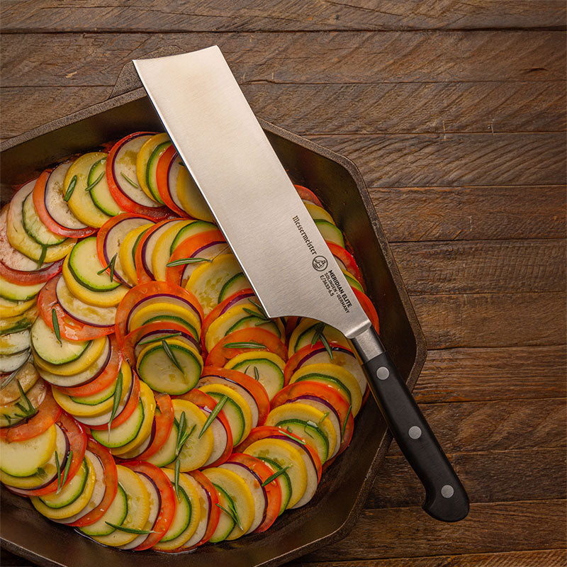 Messermeister Meridian Elite Nakiri Knife resting on an iron pan with cut vegetables in spiral formation and rosemary garnish
