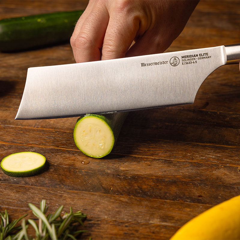 Messermeister Meridian Elite Nakiri Knife slicing zucchini on wooden cutting board with squash and rosemary garnish
