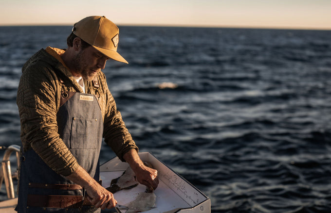 Chef Adam Glick on a boat at sunset, wearing overalls and a cap slicing fish with the Messermeister and Iron and Resin Fillet Knife.