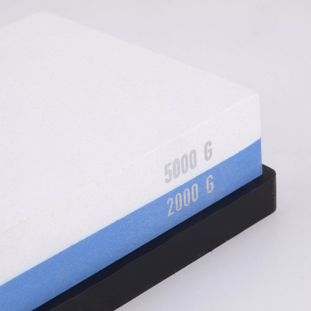 Messermeister  dual sided Polishing Stone shows the right corner/edge. The top layer is white with silver "5000 G" printed on the edge. Bottom layer is blue with silver "2000 G" printed on edge. The item sits in a black silicone base.