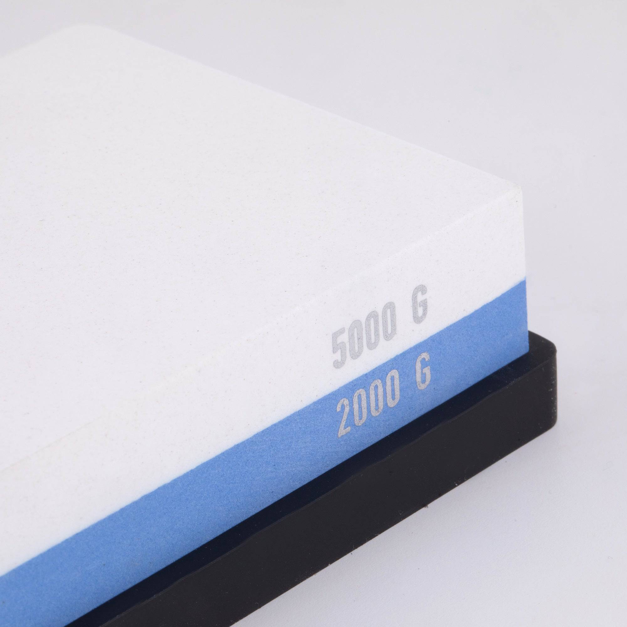 Messermeister  dual sided Polishing Stone shows the right corner/edge. The top layer is white with silver "5000 G" printed on the edge. Bottom layer is blue with silver "2000 G" printed on edge. The item sits in a black silicone base.