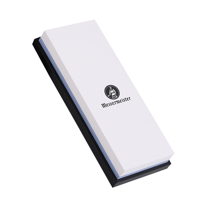 Messermeister  2000 & 5000 Grit Polishing Stone shows a dual sided rectangle 8 inch x 3 inch x 1 inch. The top layer is white with black "Messermeister" logo.  Bottom layer is blue. The item sits in a rectangular black silicone base. Image tilted up and to the left.