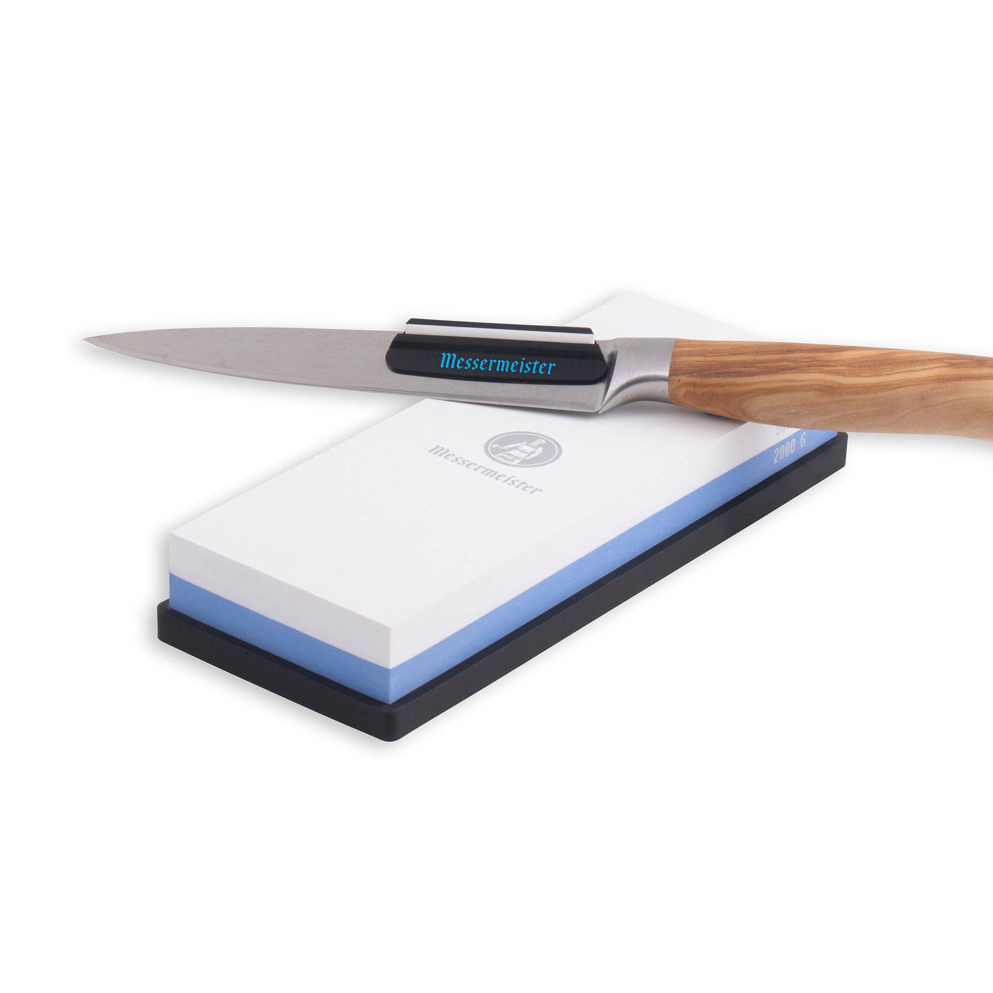 Messermeister Polishing Stone is a dual sided rectangle 8 inch x 3 inch x 1 inch. The item sits in a rectangular black silicone base. A wooden knife with angle guide is resting along the top of the stone.