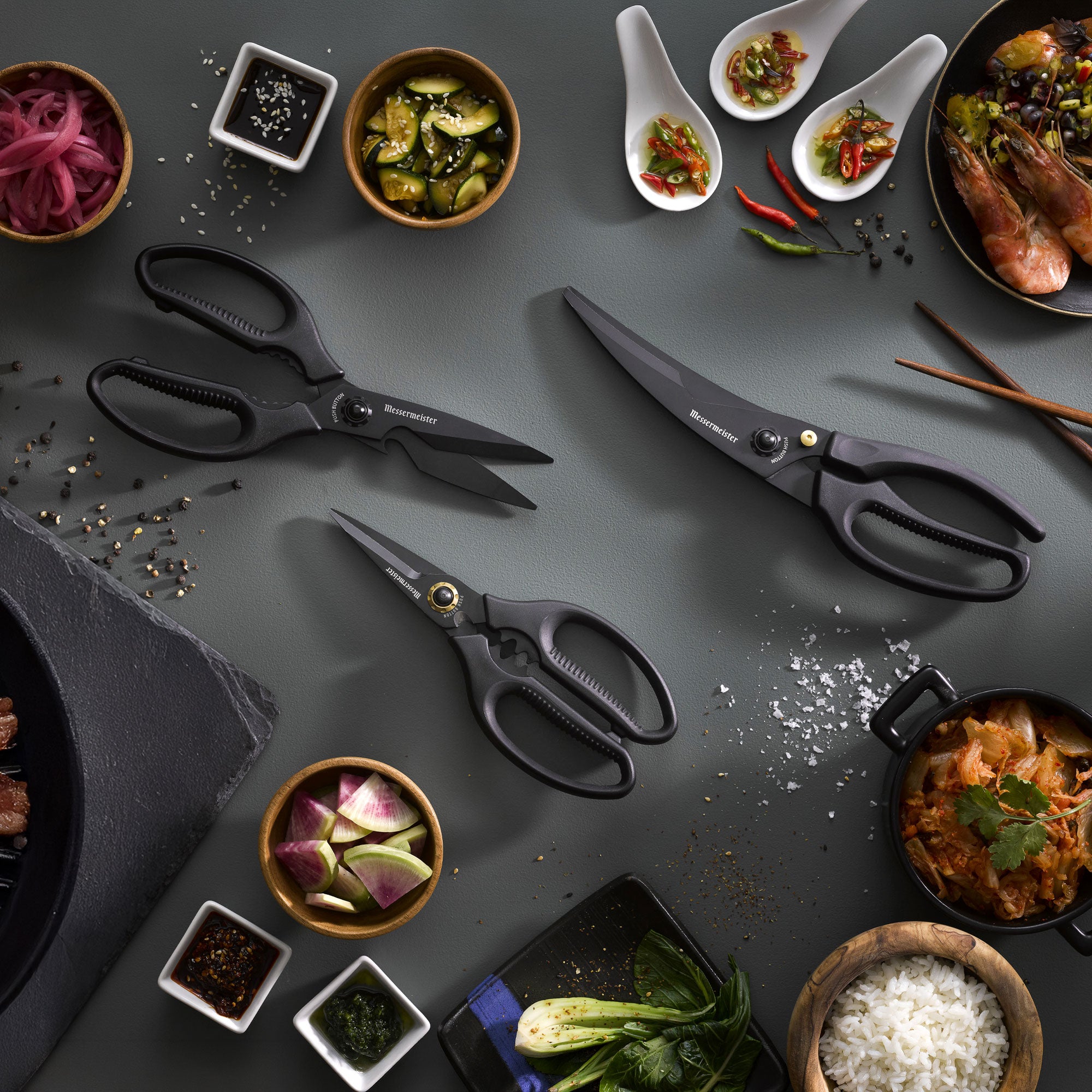 3 of the all black Messermeister Push Button Shear Collection on a dark gray surface with Korean BBQ dishes that have rice, shrimp, kimchi, pickled onions and spices