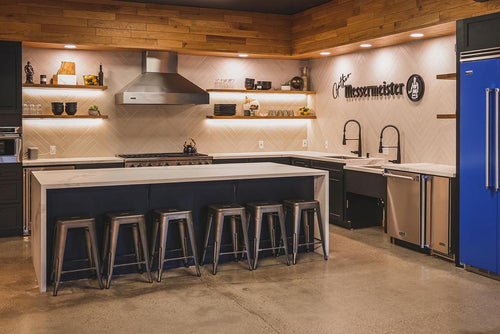 Messermeister Showroom with a kitchen that has a large island with 6 metal chairs in front of it. It has a large Viking stove and hood with floating shelves, a blue refrigerator, and a sign that says Messermeister and their logo. 