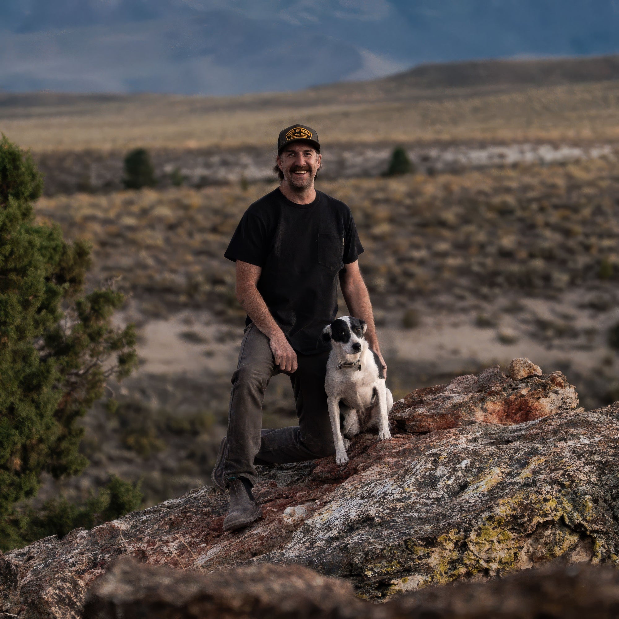 A man kneeling on a large rock in a desert landscape, wearing a black t-shirt, olive pants, and a black cap with a yellow logo, while sitting next to a black and white dog.