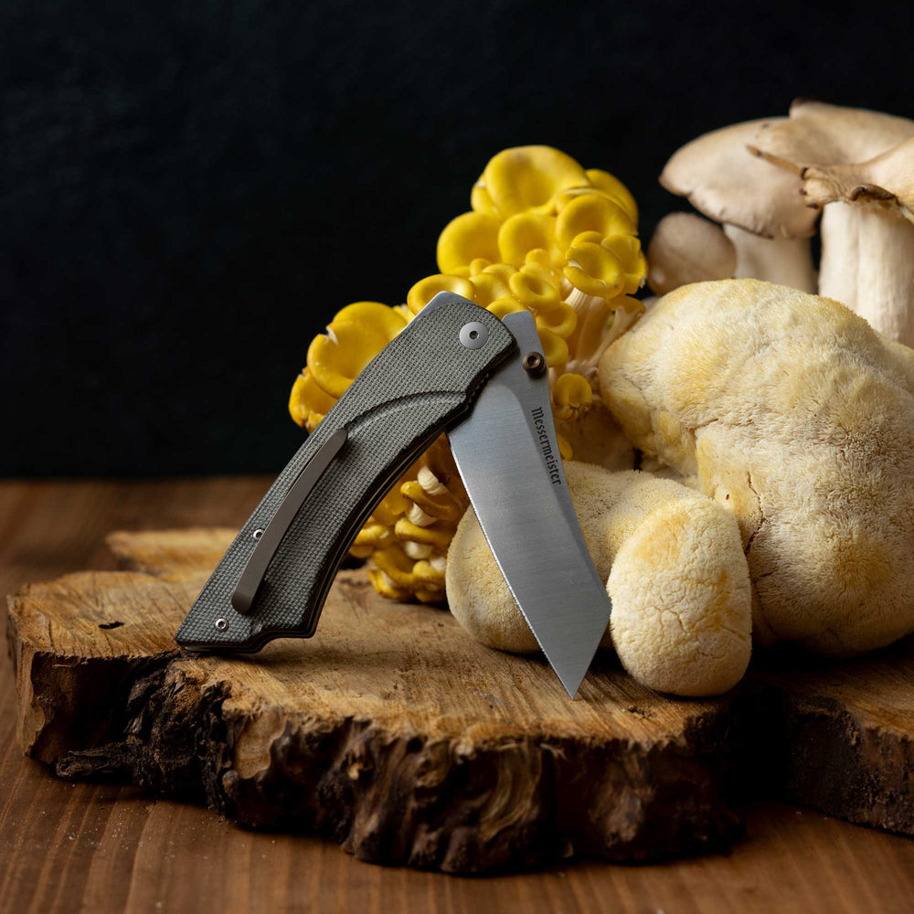 Messermeister Ojai Damasteel Folder half open on a wooden block in front of mushrooms