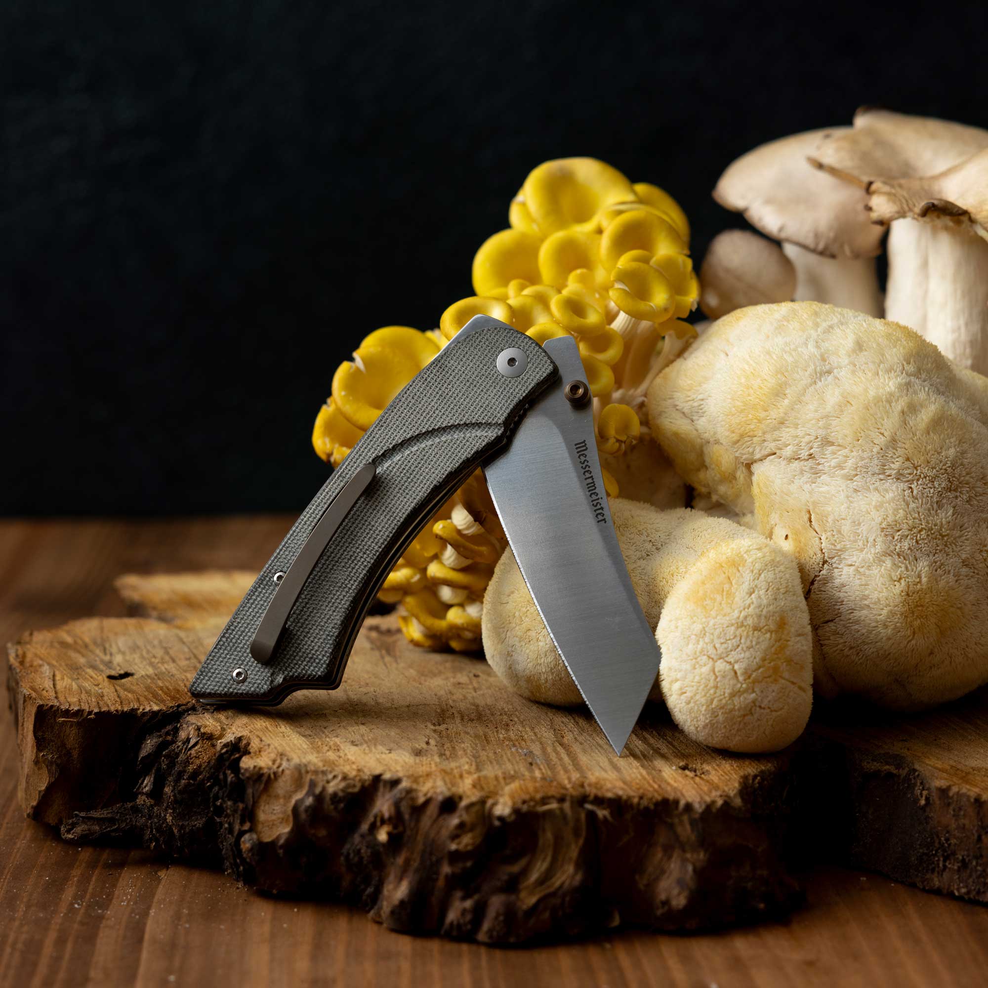 Messermeister Ojai Damasteel Folder half open on a wooden block in front of mushrooms