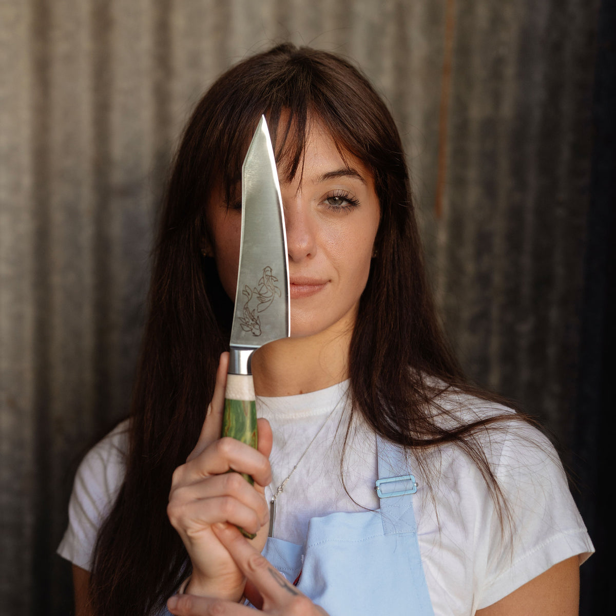 A woman with long brown hair stands in a narrow space with corrugated metal walls, holding a knife with a green-dyed wood handle. She is wearing a white t-shirt and a light blue apron.