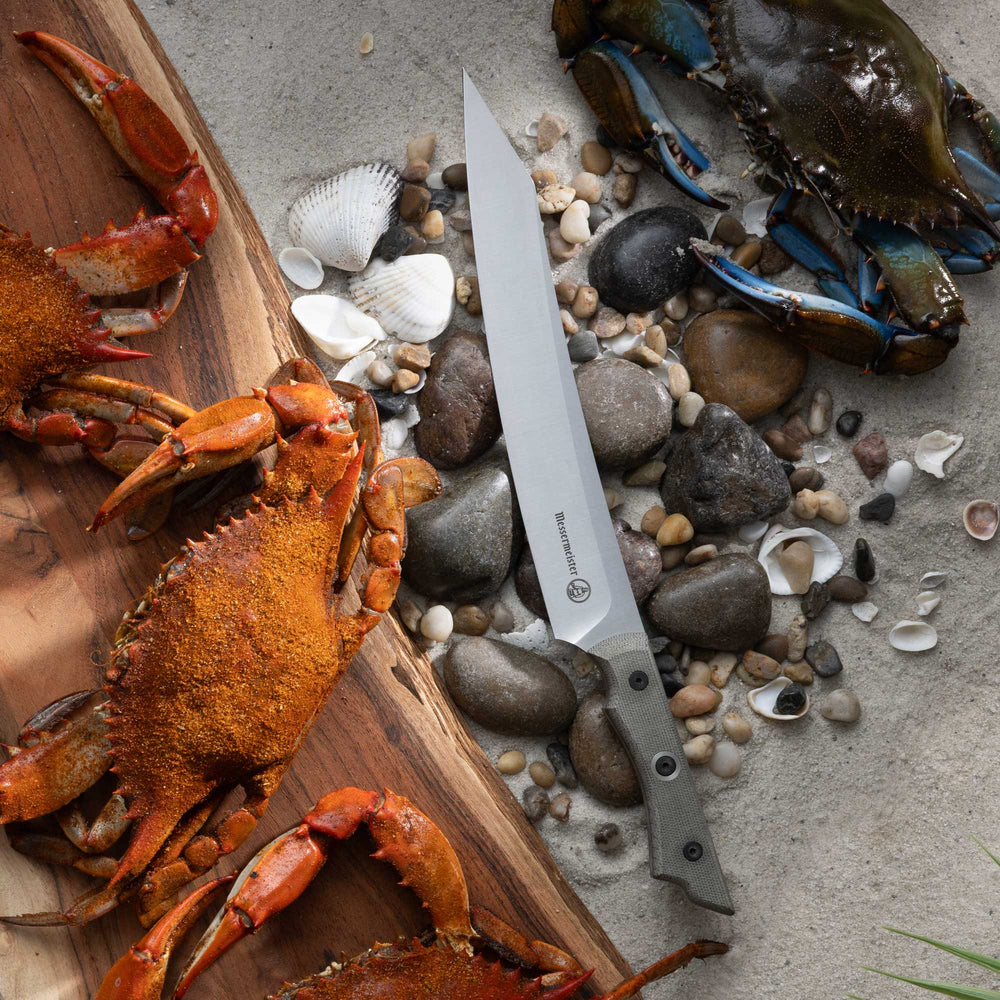 Slicing knife with a long stainless steel blade and textured light-colored handle placed among small stones and seashells, beside cooked crabs on a wooden board and a raw crab nearby