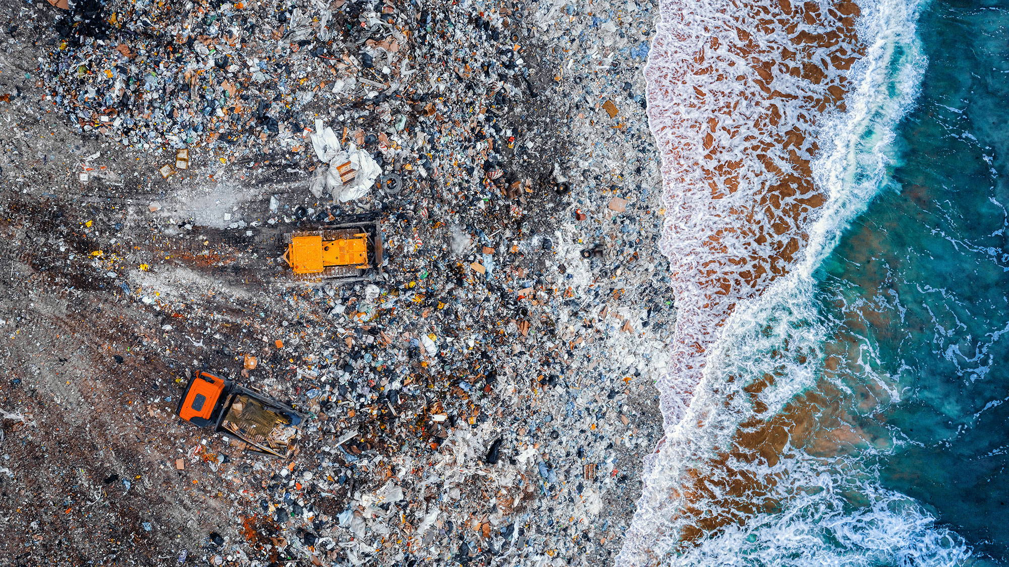 Two orange vehicles on a polluted beach with ocean waves.