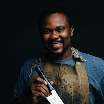 A smiling man wearing a dusty brown leather apron over a blue t-shirt, holding a knife with a blue handle and white paper label against a dark background.