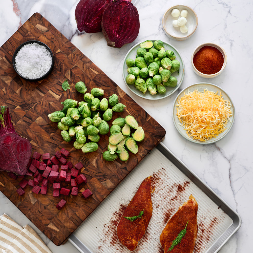 Teakhaus Prep to Pan cutting board with fresh vegetables, seasoned chicken in the slide-out pan, and bowls of cheese and seasoning nearby.