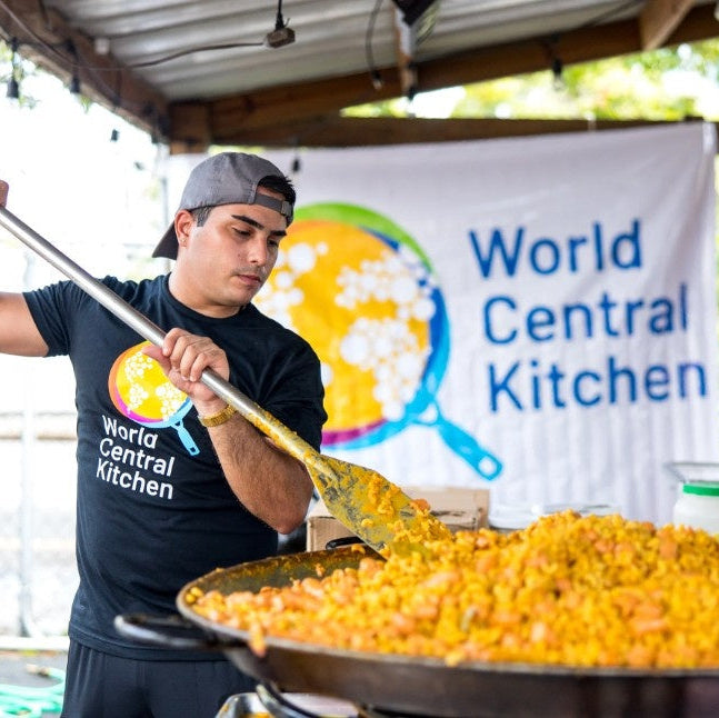 A man  with a World Central Kitchen t-shirt stirring a huge skillet of paella with a World Central Kitchen banner in the background. 