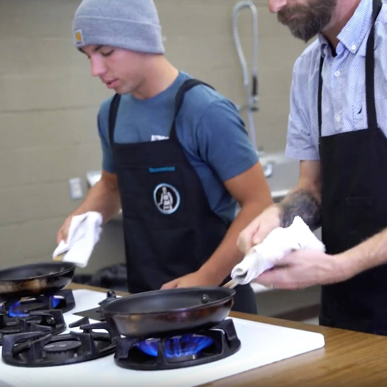 A teacher and student in aprons cooking on stovetops in a kitchen setting.