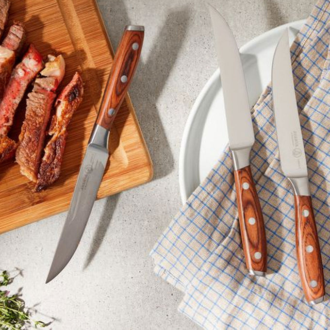 Knives with wooden handles on a checkered cloth next to a cutting board with cooked meat.