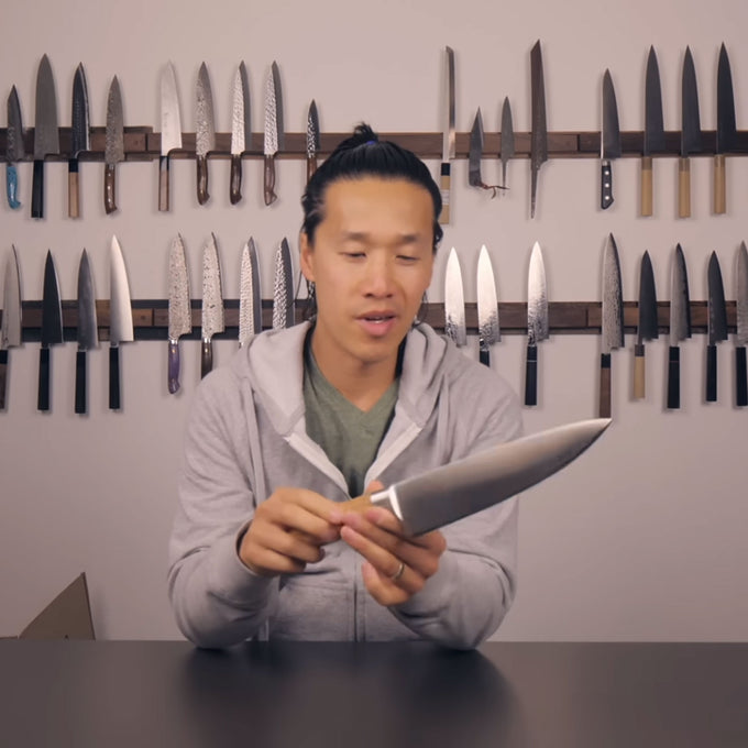 Man holding a Messermeister Oliva Chef knife in front of a wall of knives