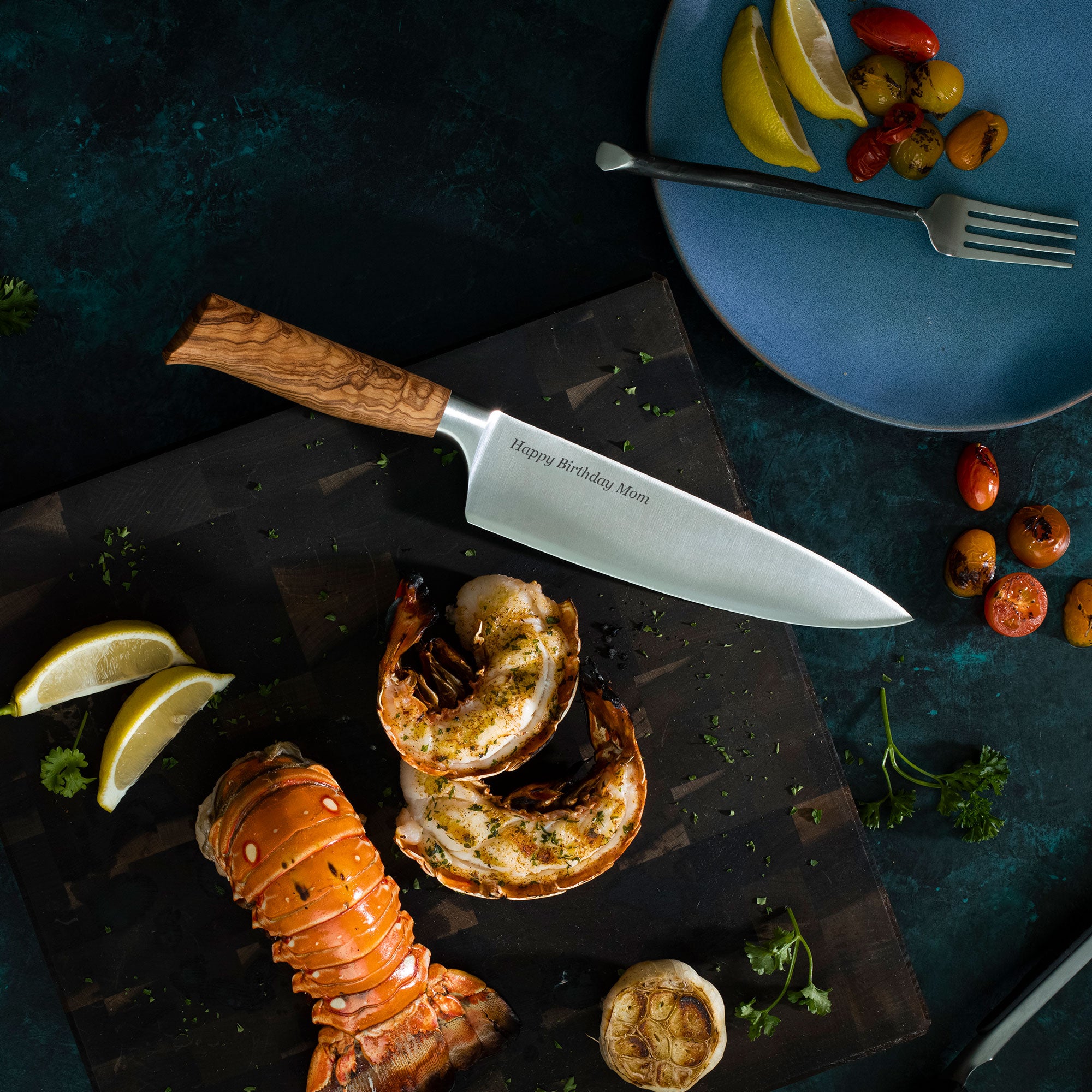 Lobster tail and shell on a dark surface with a knife, lemon wedges, and a plate of vegetables.