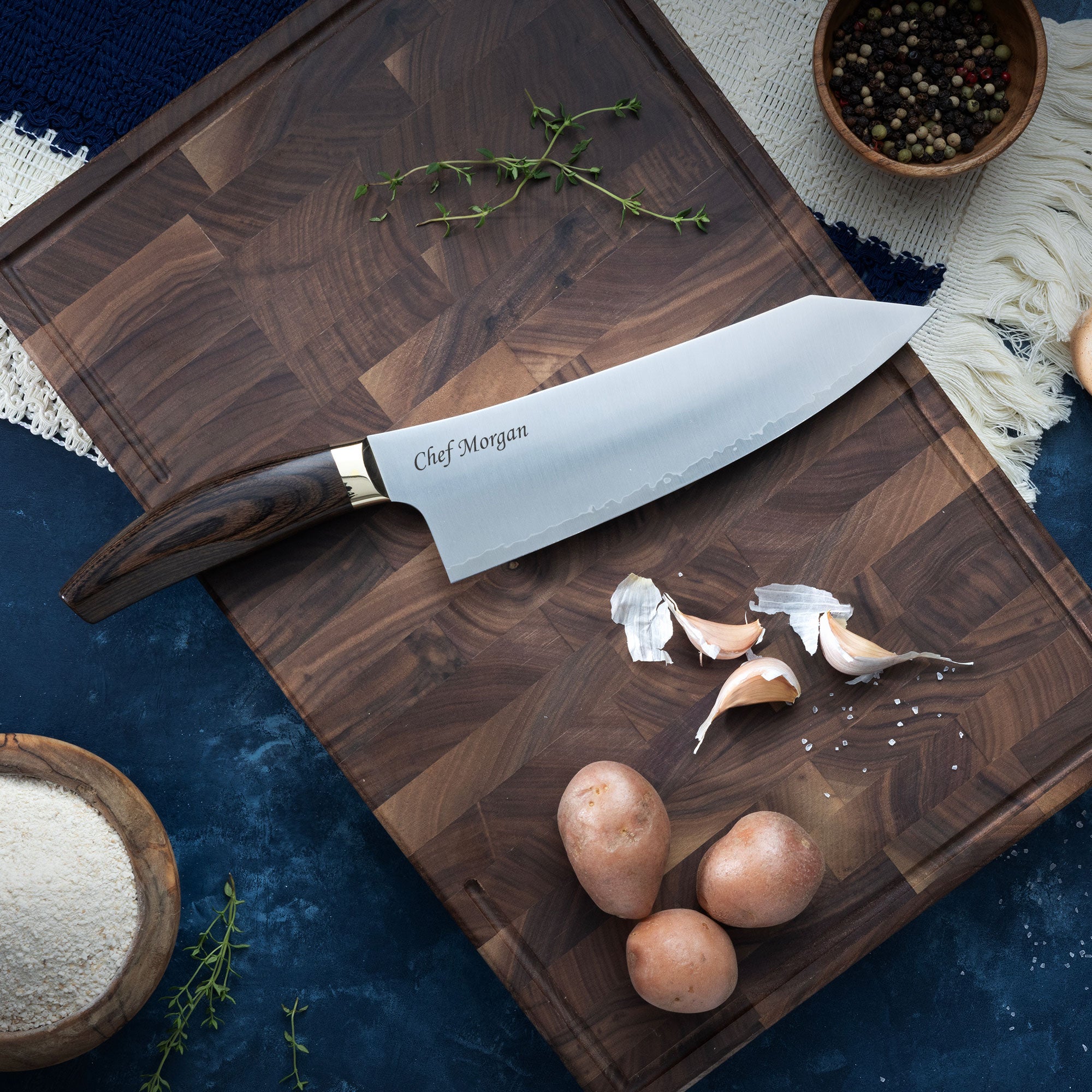 Chef's knife on a wooden cutting board with garlic and herbs, on a dark blue surface.