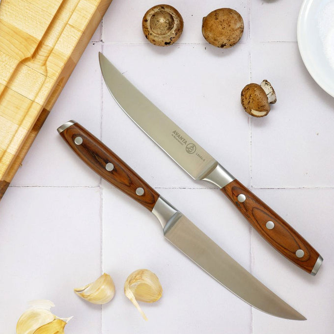 Two Avanta steak knives with wooden handles on a white surface with garlic and mushrooms.