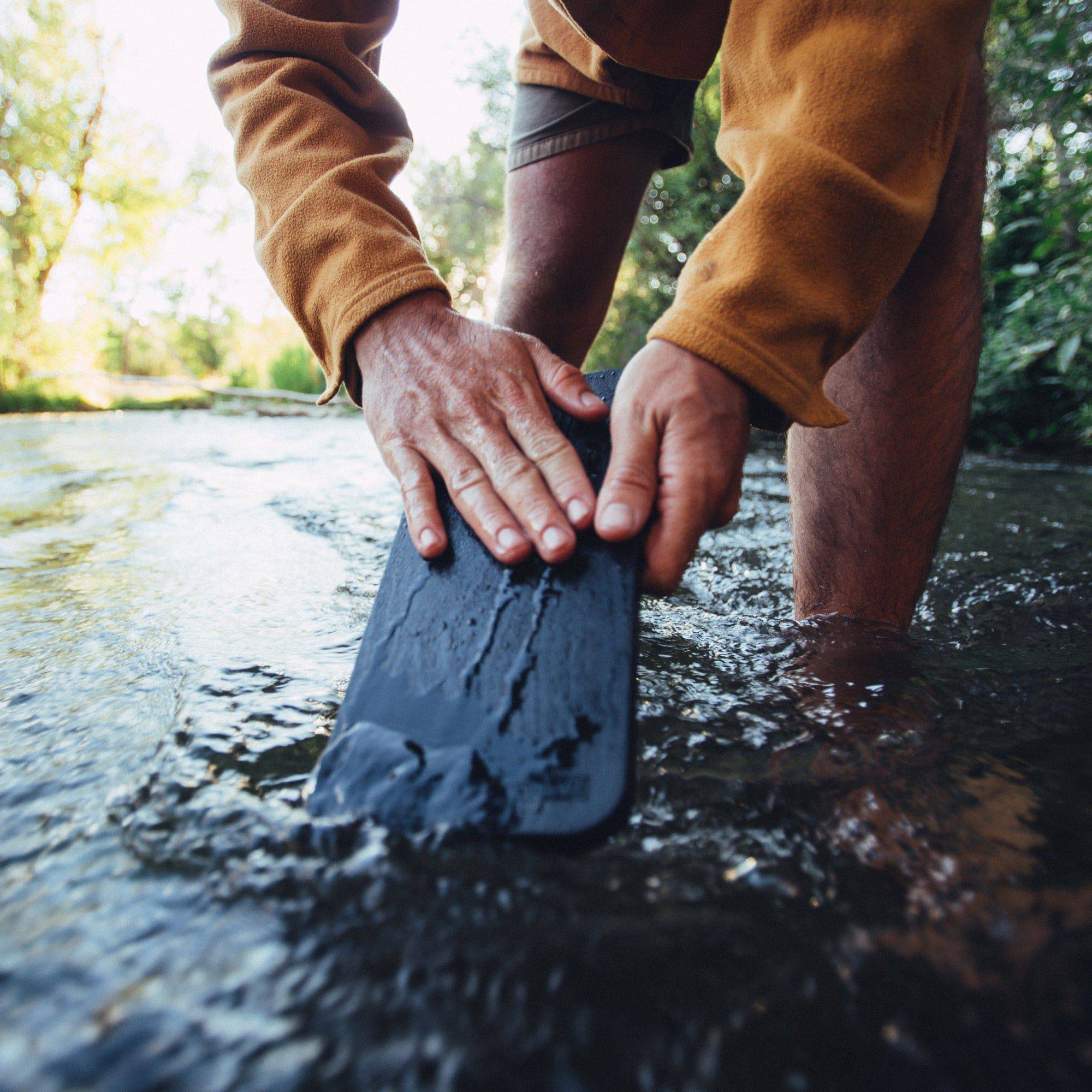 Close up photo of person rinsing off the Messermeister Adventure Chef Folding Faux Wood Cutting Board  in a creek with trees in the background