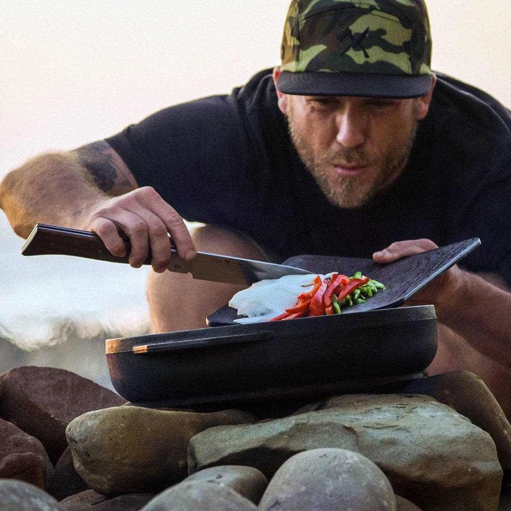 Photo of person outside using the Adventure Chef 6 inch Chef's knife to ease cut food off of cutting board into iron skillet that is resting on fire pit