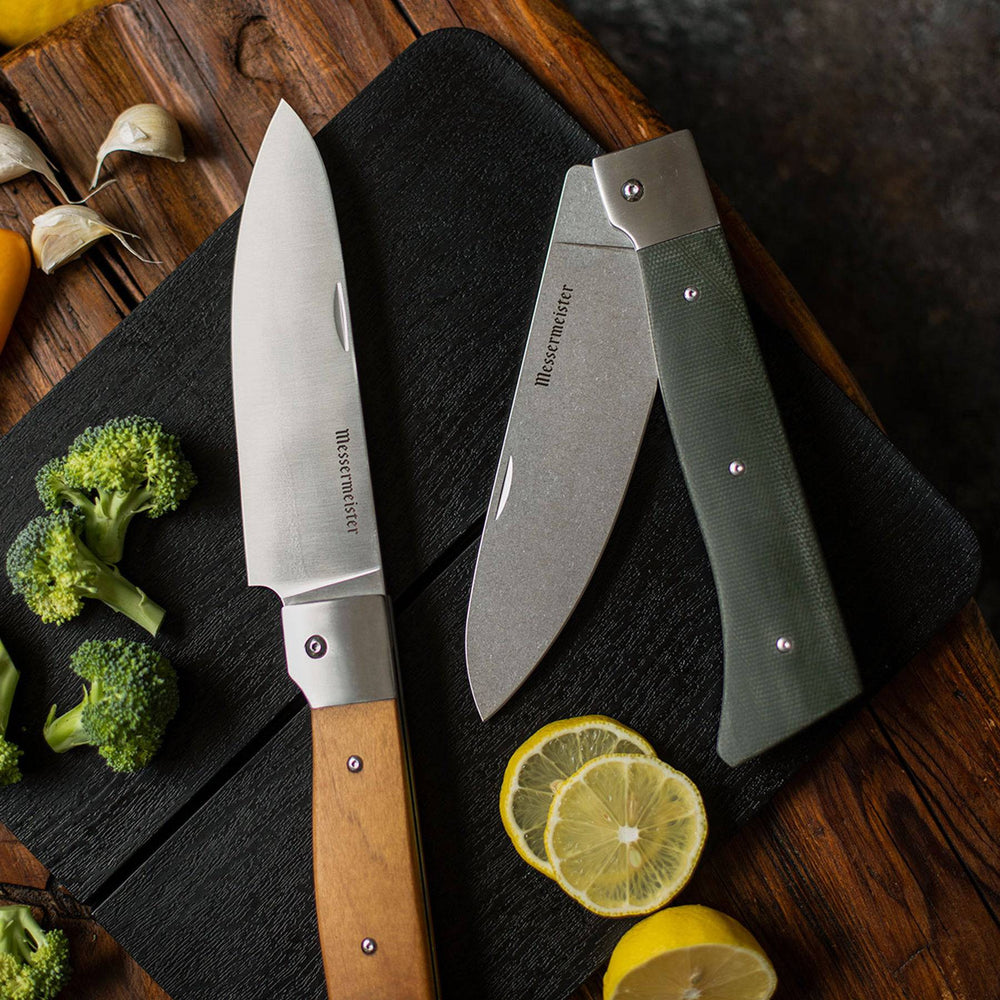 Messermeister Adventure Chef Folding 6 inch Chef's Knife with Maple handle shown fully extended lying on the Adventure Chef folding cutting board next to the Linen chef's knife with sliced fruits and vegetables