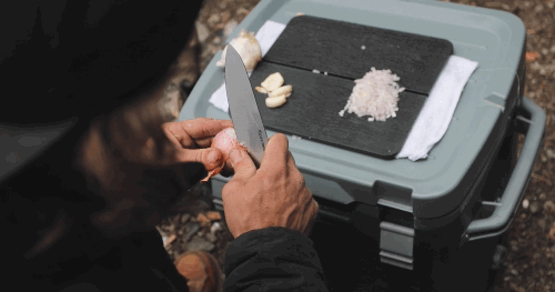Video of a person at a campsite using the Adventure Chef 6 inch Chef's Knife to peel a shallot with cut vegetables on a cutting board sitting on top of a cooler