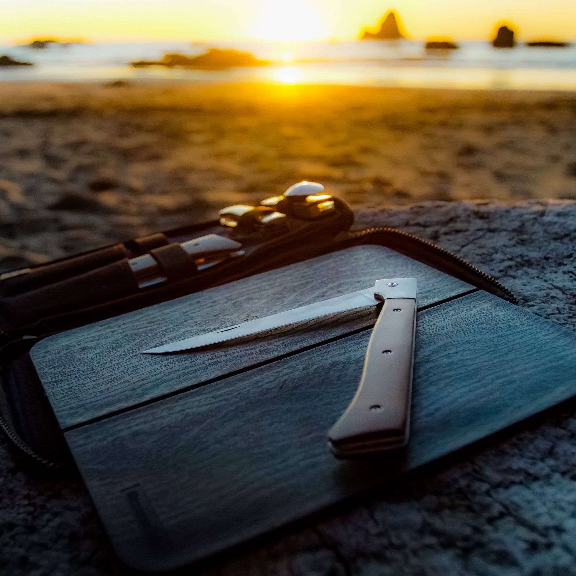 Photo at the beach with the sunset and water in the background and the folding 6 inch fillet knife resting on the faux wooden cutting board in the foreground