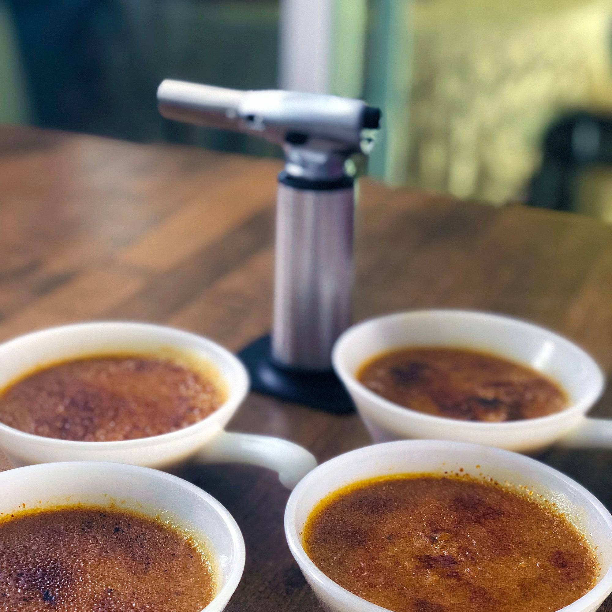 Messermeister Cheflamme Culinary Torch displayed beside four freshly prepared crème brûlée bowls with caramelized golden sugar tops, showcasing professional dessert preparation, gourmet presentation, and precision culinary torch performance