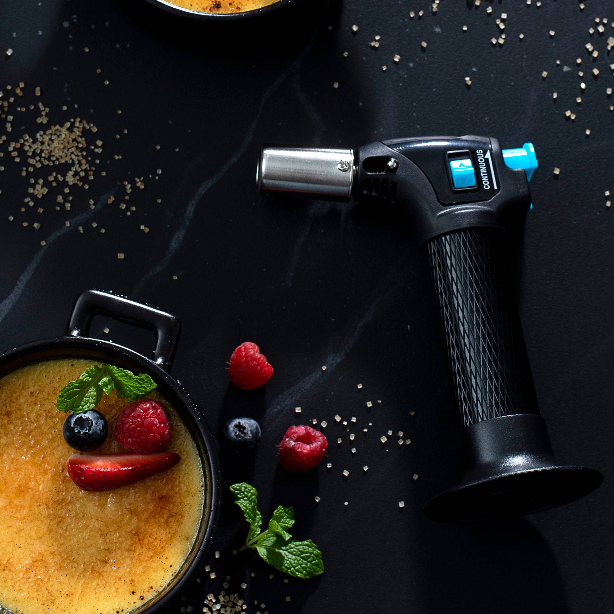 Photo of the Messermeister Cheflamme Mini Culinary Torch with black handle and silver nozzle laying next to a pot of creme brulee and berries