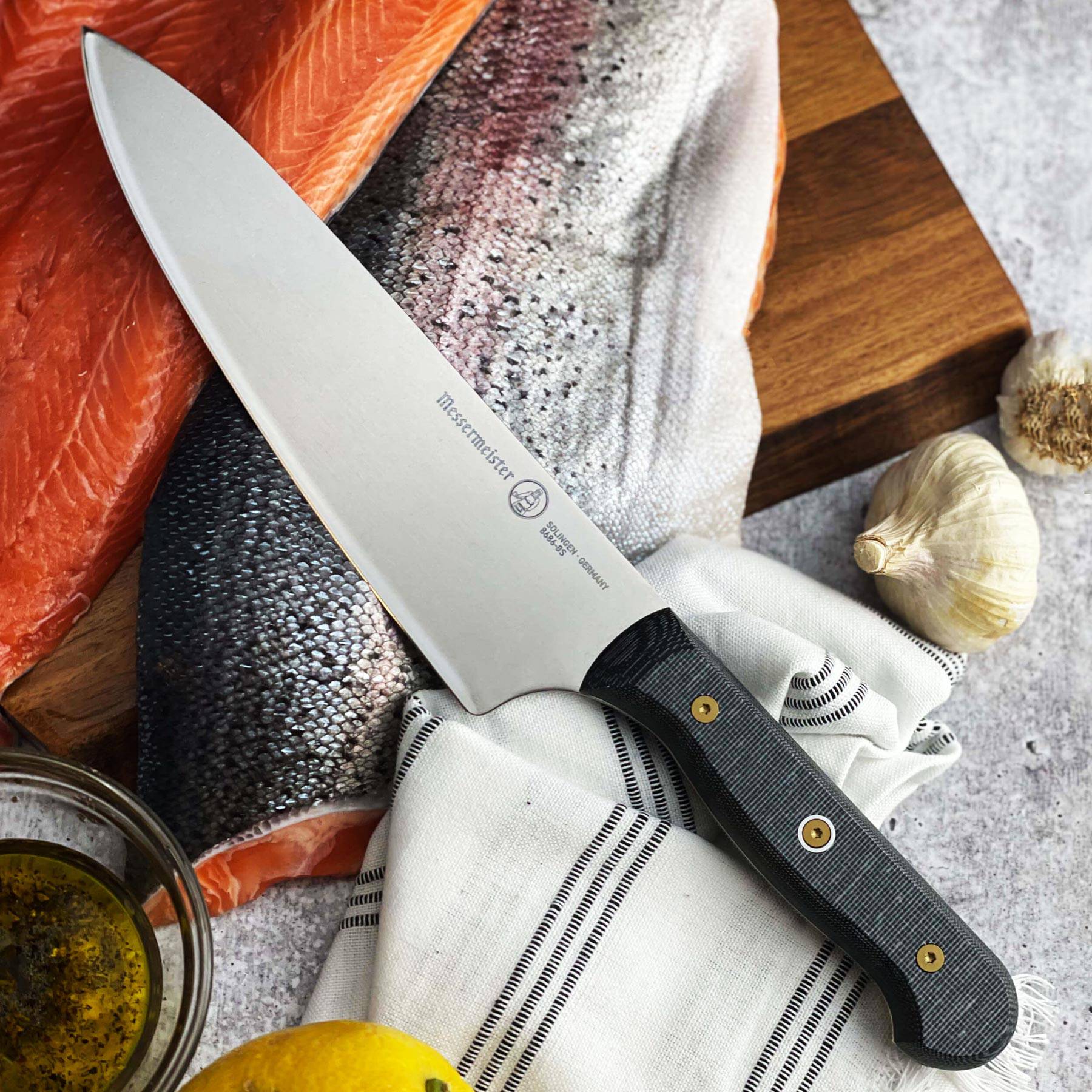 Messermeister Custom Chef’s knife on raw salmon fillet with garlic cloves and oil for food preparation
