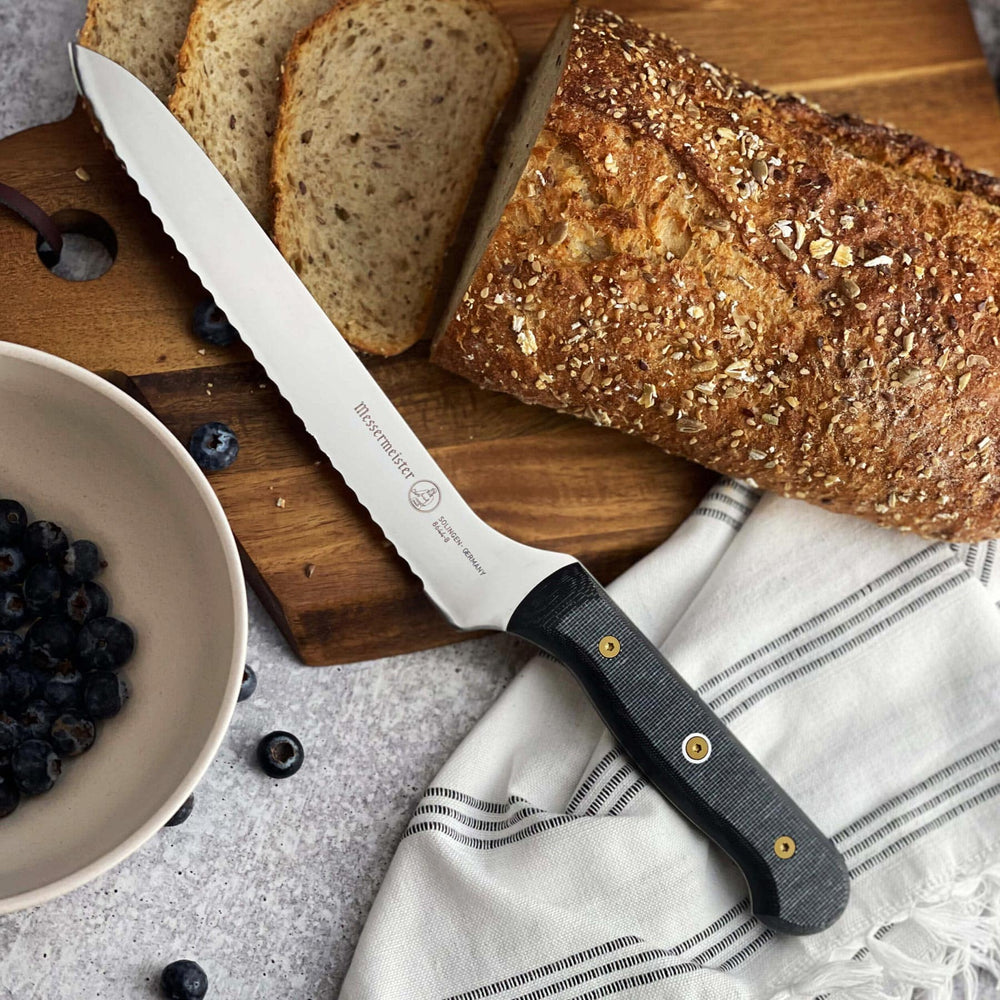Messermeister Custom Offset Scalloped Bread Knife - 8" resting on a wood cutting board with a loaf of bread and a bowl of blueberries
