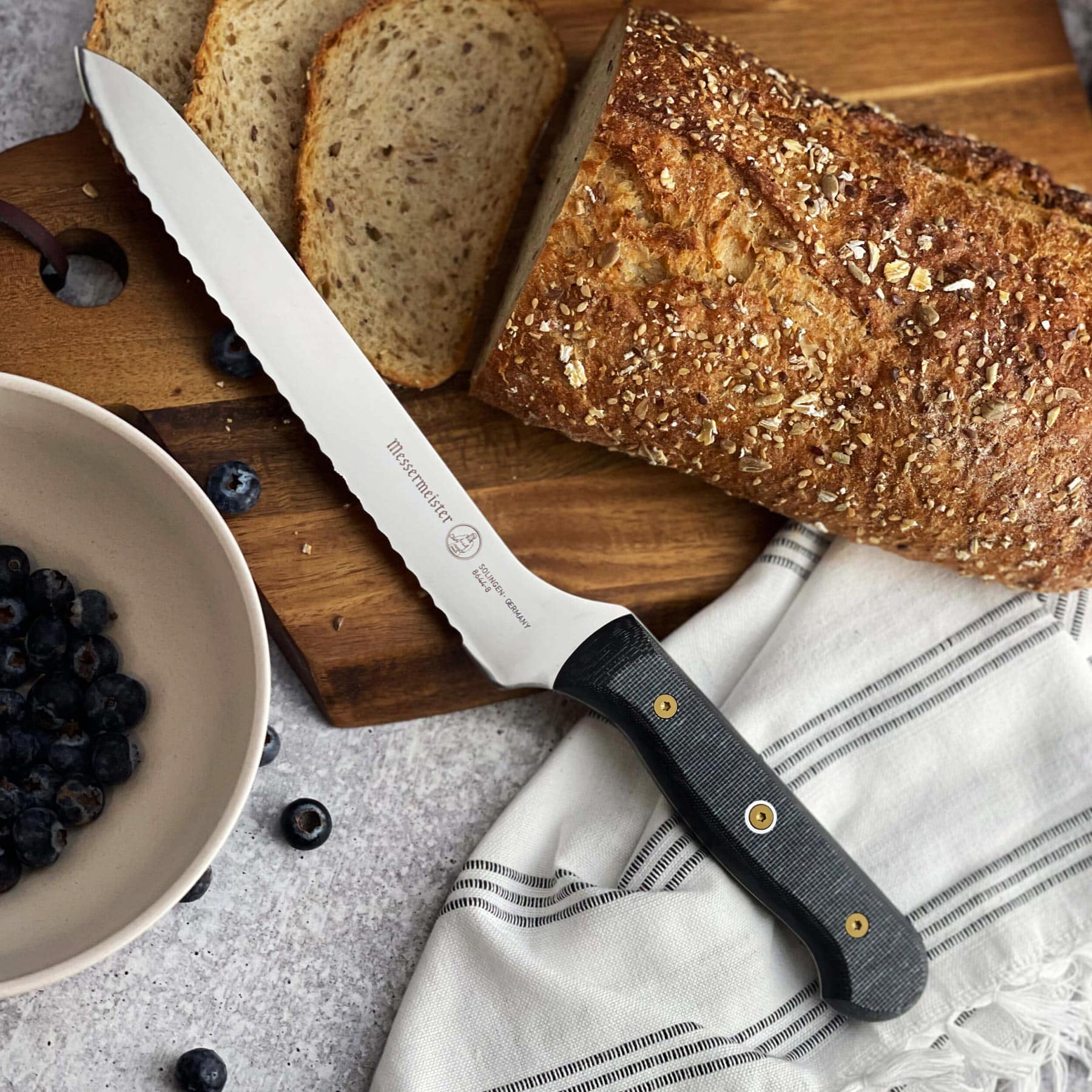 Messermeister Custom Offset Scalloped Bread Knife - 8" resting on a wood cutting board with a loaf of bread and a bowl of blueberries
