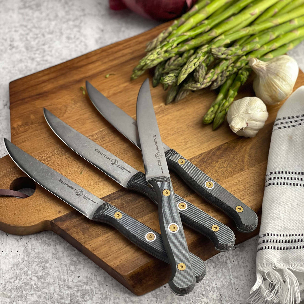 Four-piece Messermeister Custom steak knife set with 5-inch blades arranged on a wooden cutting board beside fresh asparagus and garlic, featuring stainless steel blades and ergonomic modular handles with textured linen micarta scales, highlighting sharp edges