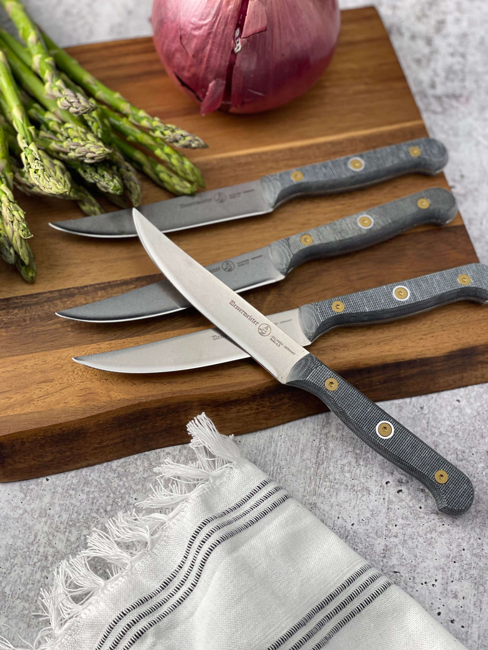 Messermeister Custom steak knife set with 5-inch blades arranged on a wooden cutting board beside fresh asparagus and onion, featuring stainless steel blades and ergonomic modular handles with textured linen micarta handle