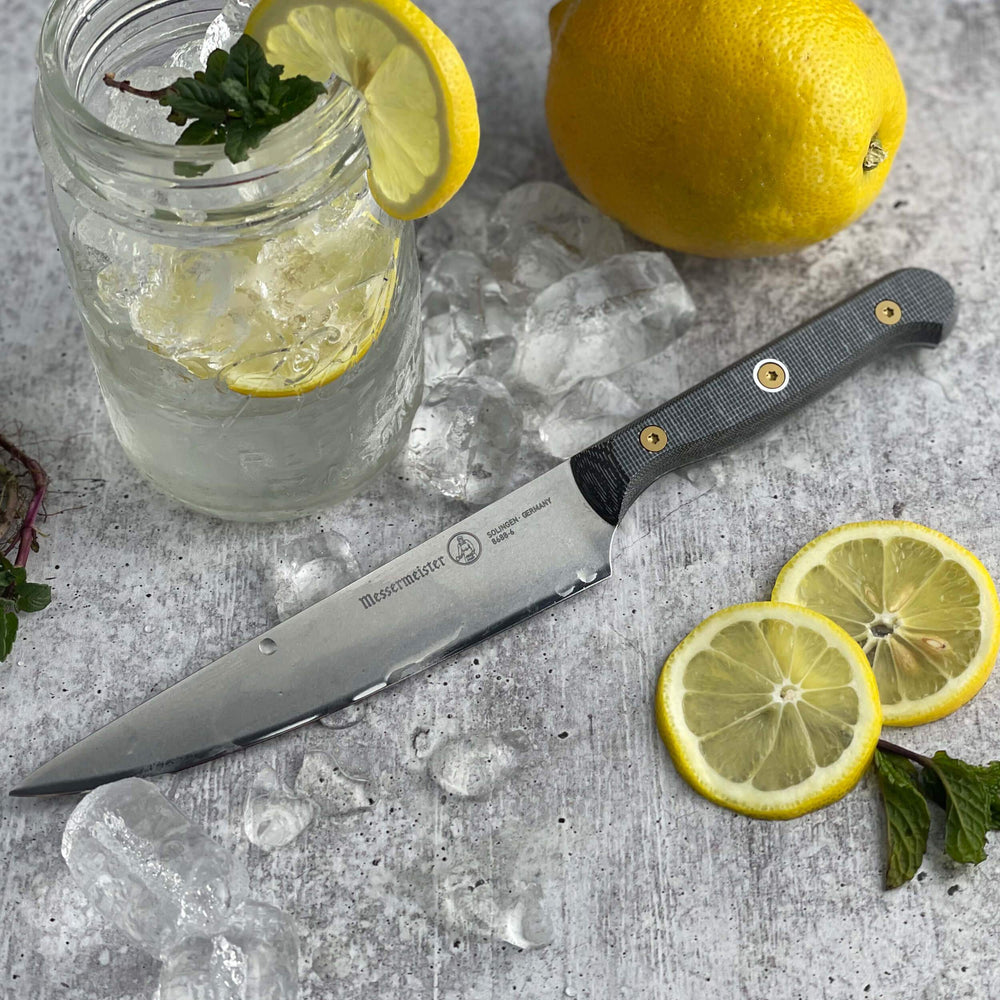  Messermeister Custom 6-inch utility knife resting on gray surface surrounded by fresh lemons and mason jar of lemon water