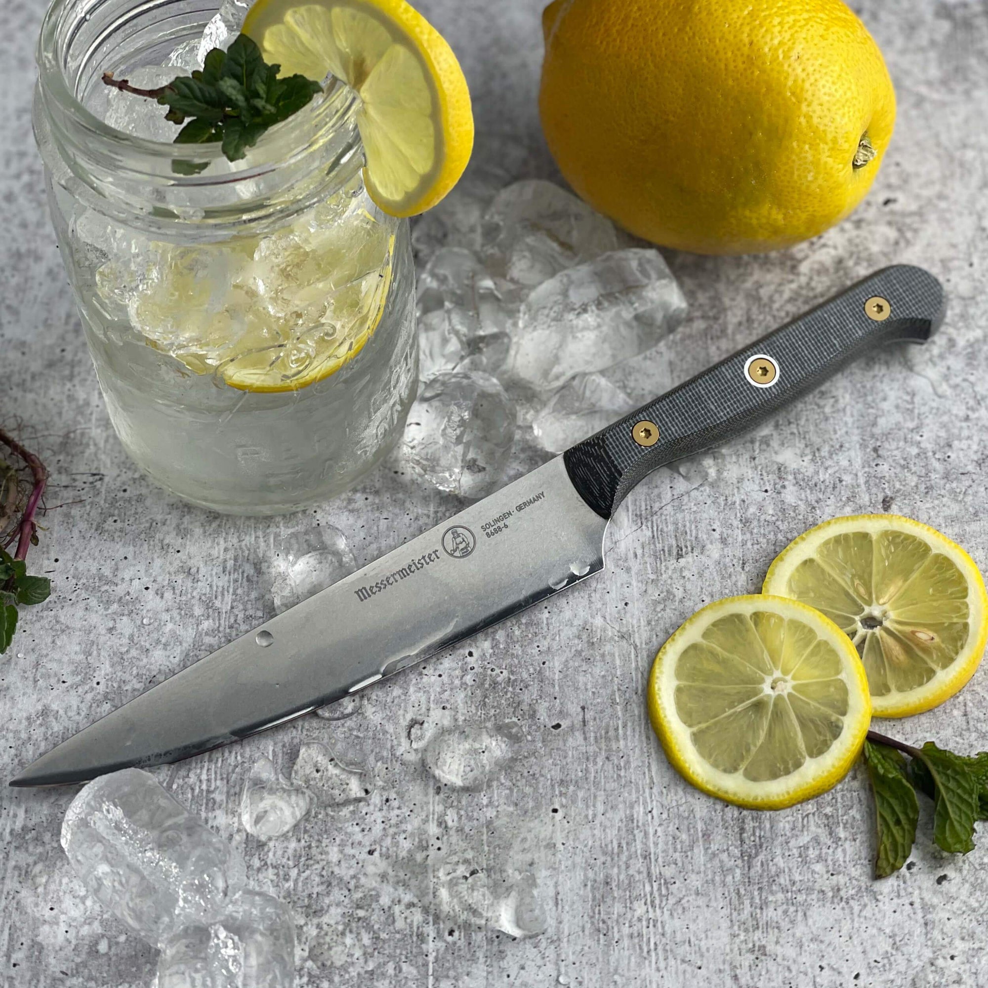 Messermeister Custom 6-inch utility knife resting on gray surface surrounded by fresh lemons and mason jar of lemon water